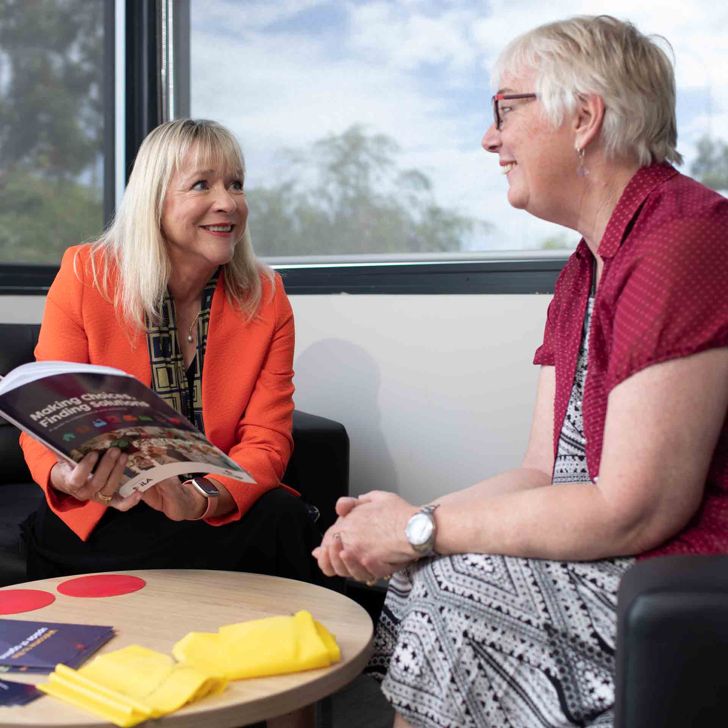 Two people indoors near large windows, one showing brochure at table 