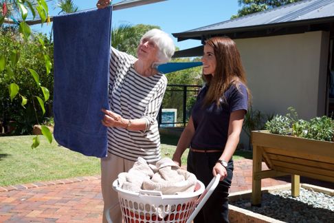 Older woman hanging towel on clothesline with support worker holding laundry basket
