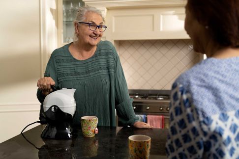 An older woman using a tipping kettle talking with her friend in a kitchen