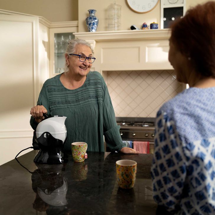 An older woman using a tipping kettle talking with her friend in a kitchen