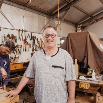 A senior man smiling into camera standing in a woodworking shed.