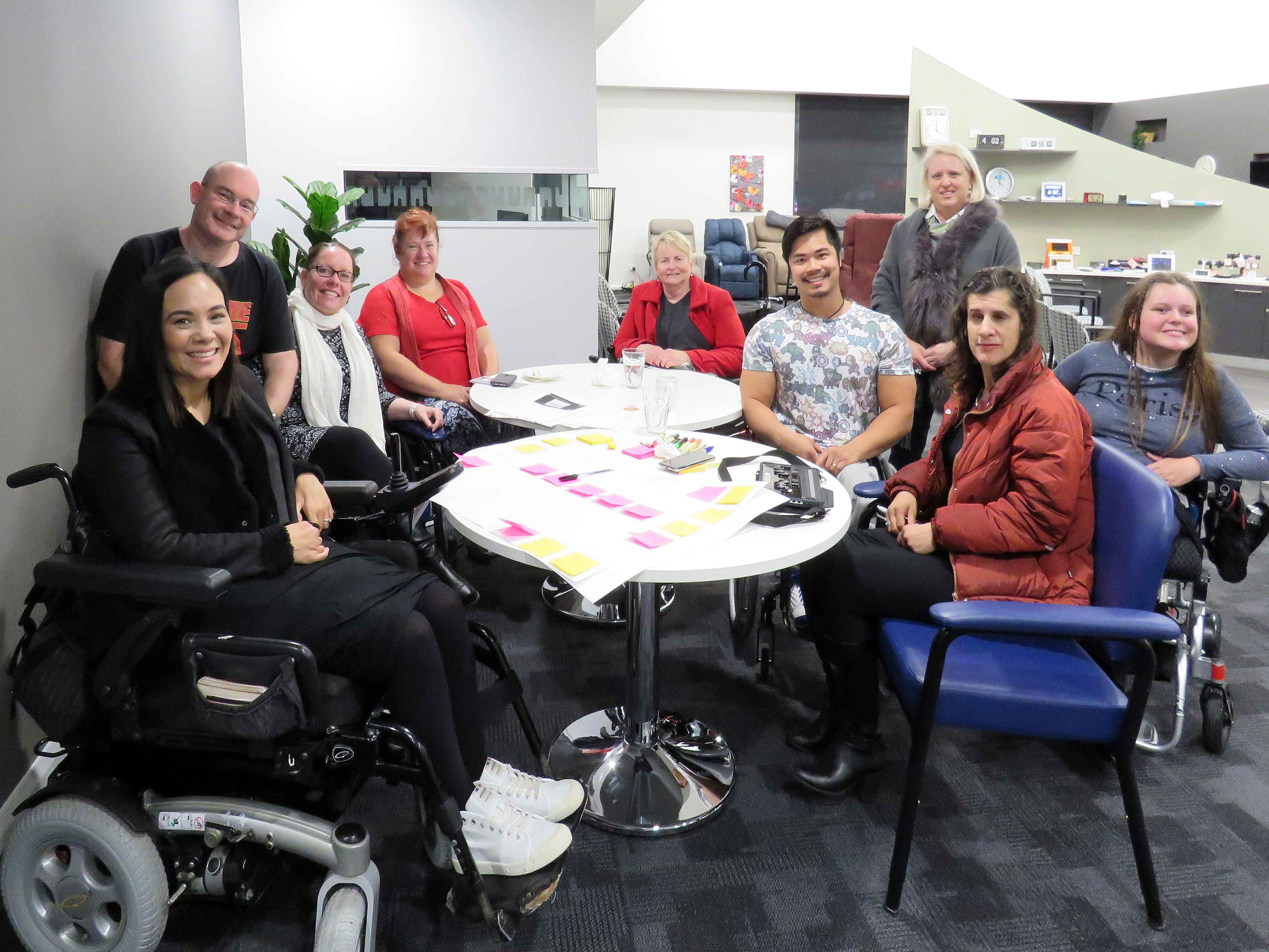A group of people with disability are seated around a table with papers and pens in front of them.
