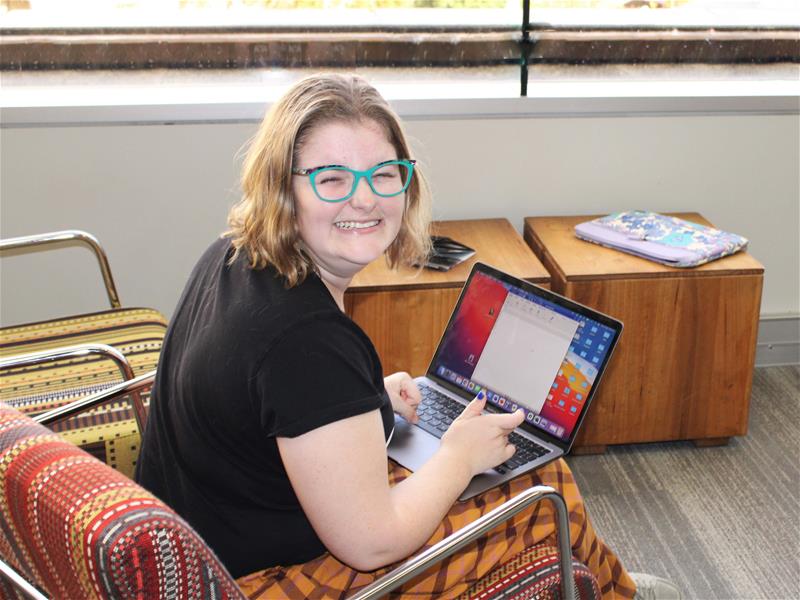 A young lady is smiling at the camera as she sits with a laptop open on her lap