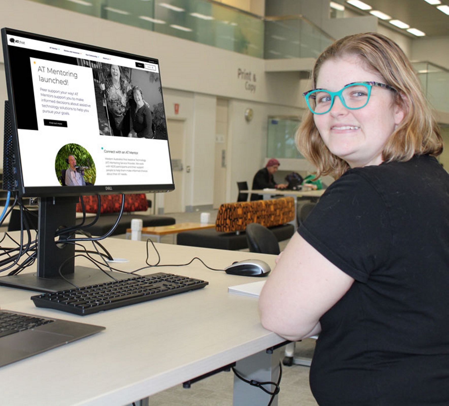 A young lady is seated at her computer. She is turned to smile at the camera. On her screen is the AT Chat website.