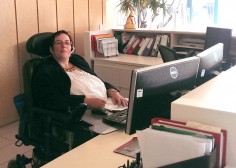 female wearing glasses and a headset in wheelchair at a desk with two computer screens in an office