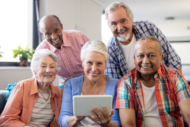 smiling-senior-people-with-digital-tablet- Five older people smile at the camera one holds a tablet
